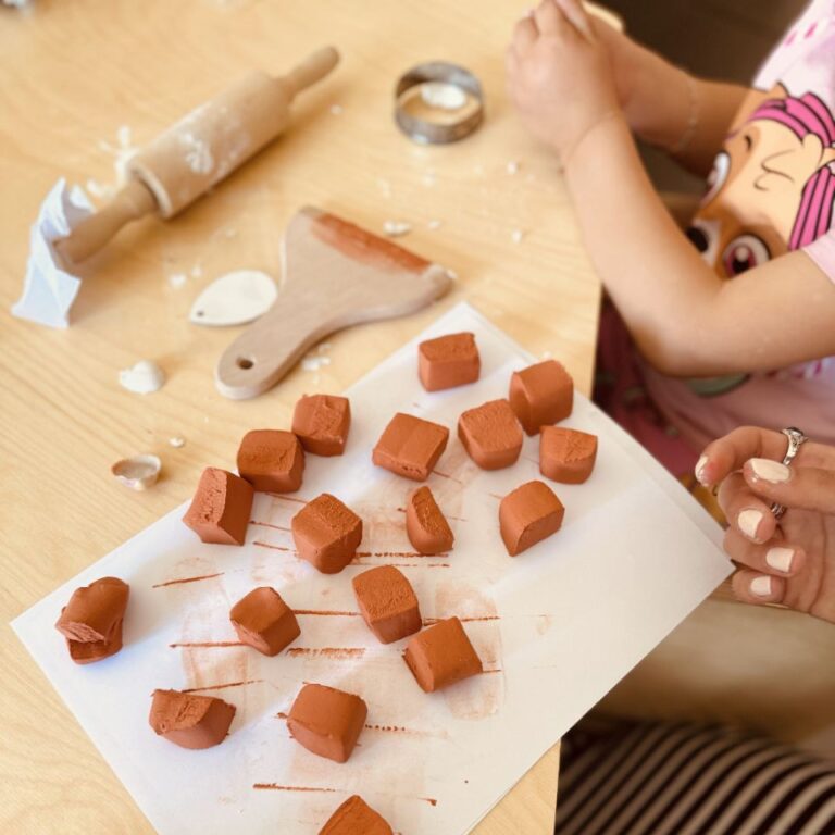 Foto di un laboratorio creativo di ceramica per bambini della prima infanzia dove si vedono dei pezzetti di ceramica rossa su un foglio bianco e si intravedono le mani di una bimba mentre modella la pasta
