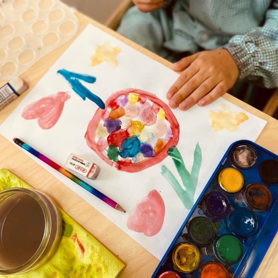Laboratorio di illustrazione: foto dall'alto di un foglio con un disegno acquerellato, acquerelli, e la manina di un bambino sul disegno